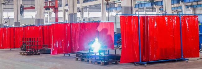 Large industrial workshop with multiple welding stations separated by red PVC welding curtains, with a welder working behind one partition.