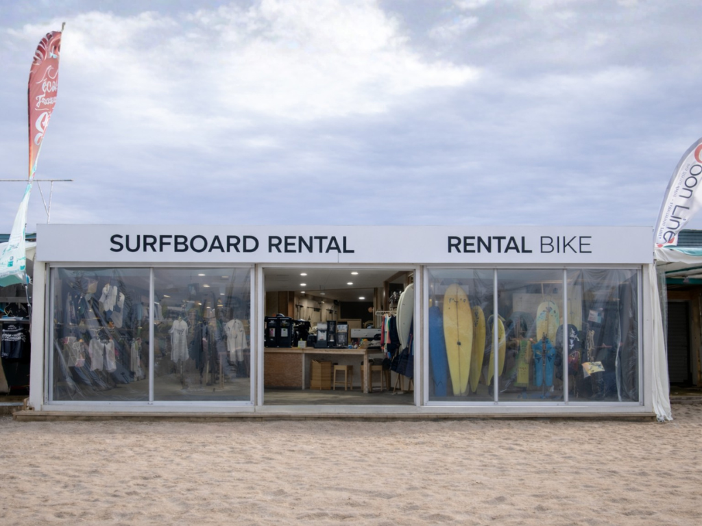 A beachside rental shop displays surfboards and clothing behind large, clear protective screens.