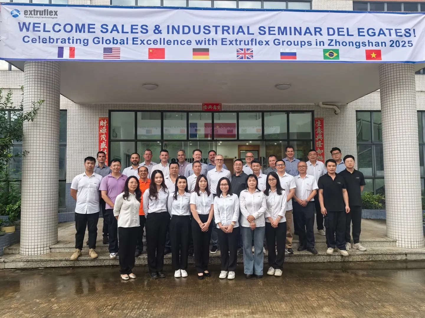 A group of Extruflex employees and international delegates pose in front of a factory building beneath a banner welcoming participants to the 2025 sales and industrial seminar in Zhongshan.