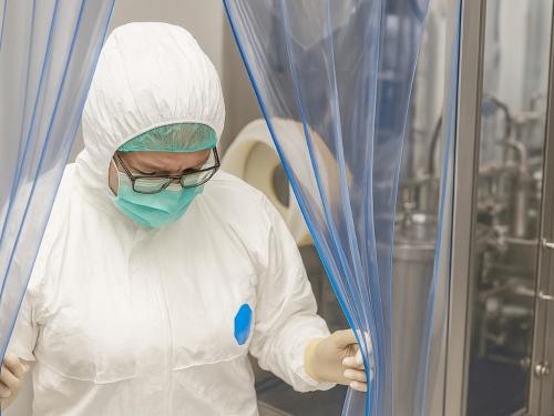 A person in a white protective suit, including a hood, gloves, and a face mask, pushes aside a blue PVC strip curtain to exit a sterile room.