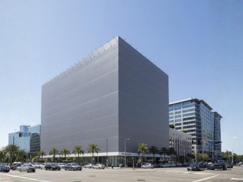 A large, square commercial building in a city covered entirely in a grey mesh or fabric facade wrap, with palm trees and street traffic visible below.