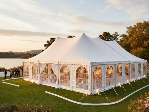 A sophisticated white pole tent with arched clear windows and white curtains set up for a wedding reception on a manicured lawn next to a lake at sunset.