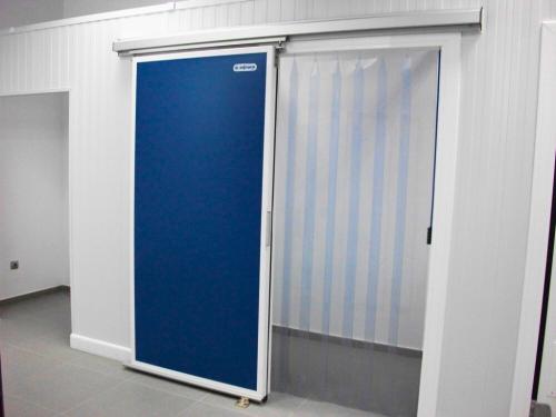 A sliding cold storage door that is half-open. The solid blue part of the door is slid to the left, revealing clear plastic strip curtains hanging in the open doorway.