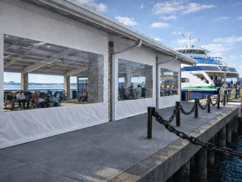 People sit in a covered waiting area on a pier next to a docked ferry, with large clear plastic windows built into white vinyl curtains.