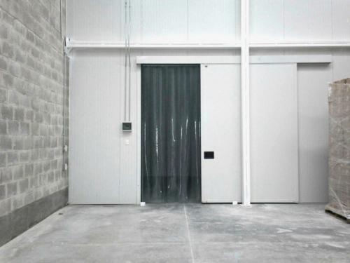 A wide shot of a warehouse with a large gray brick wall on the left. In the center are two white cold storage doors, one of which is a sliding door with clear plastic strip curtains hanging inside the opening.