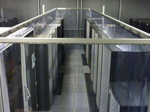 An overhead view of a data center aisle with two rows of tall, dark server racks. The aisle is enclosed by clear plastic panels and a metal frame, creating a hot or cold aisle containment system.