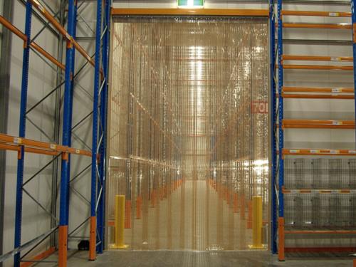  A wide-angle interior view of a warehouse with tall blue and orange metal shelving racks on either side. In the center, a walkway is covered by a plastic chain link curtain.