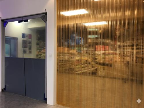 An interior view of a building with two different doorways. On the left is a door with a clear top half and a solid blue bottom half. On the right, a wide doorway is covered with translucent golden-colored plastic strip curtains. The interior of a supermarket is visible through the curtains.