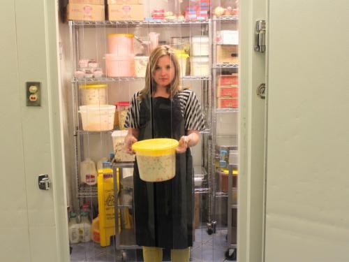 A woman in an apron stands inside a walk-in cooler, holding a large yellow lidded tub of food. Clear plastic strip curtains hang in the doorway in front of her.